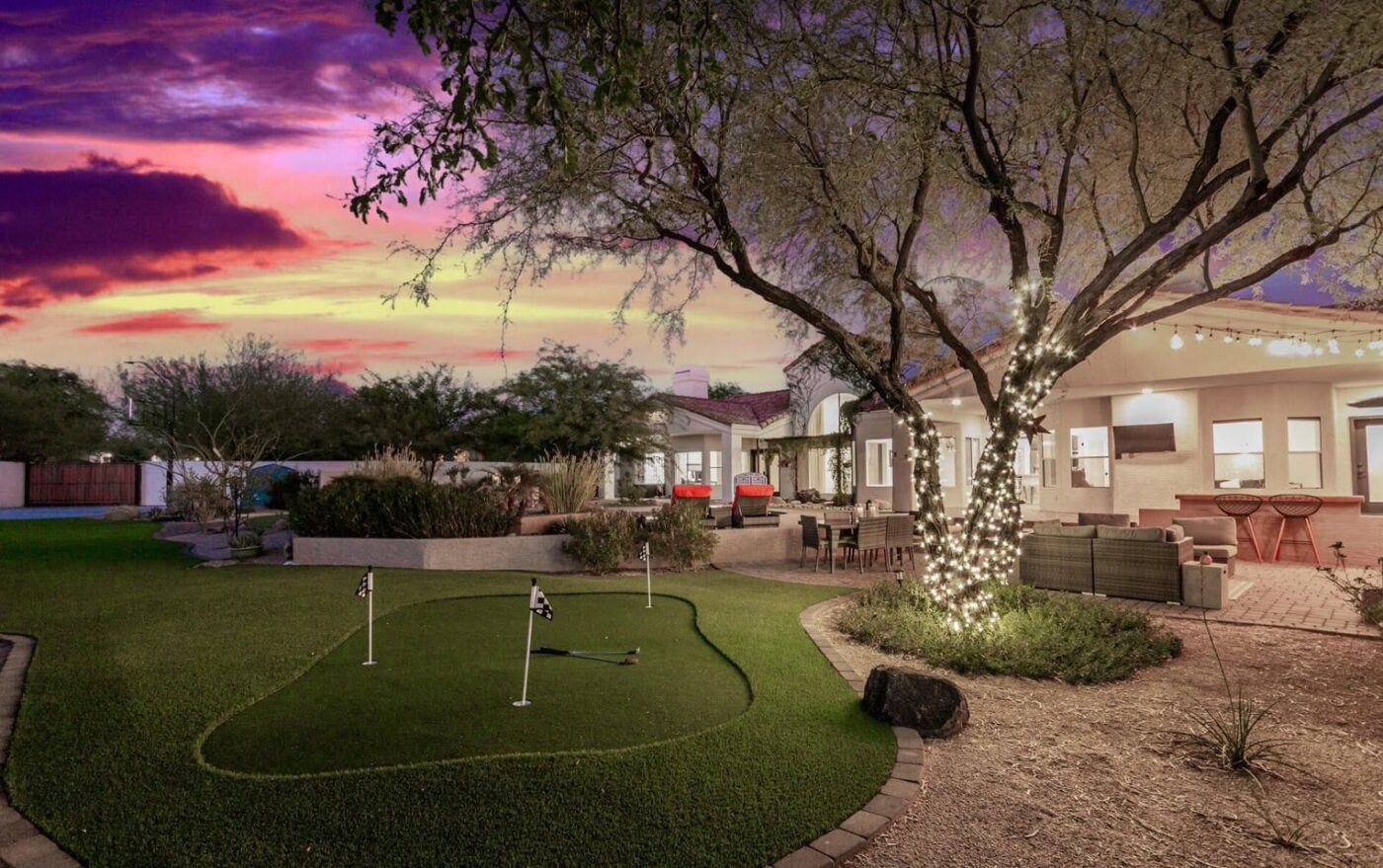 Backyard at sunset with a custom Backyard Putting Green, cozy seating area, and a tree wrapped in string lights. The patio features outdoor furniture and glowing lights beneath a vibrant purple, orange, and pink sky.
