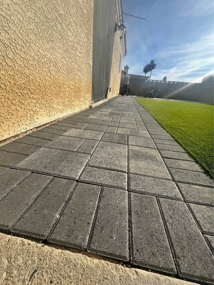 A low-angle view of a paved walkway with rectangular gray bricks beside a house, bordered by a bright green Prescott Turf and Pavers artificial lawn under a clear blue sky.