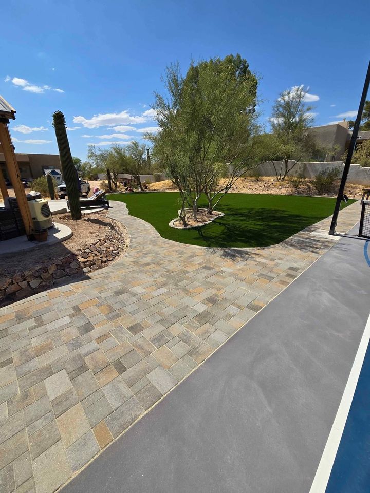 A backyard with a paved stone patio by Prescott Turf and Pavers, an artificial grass lawn, several desert trees and cacti, and a pool edge visible in the foreground under a clear blue sky.