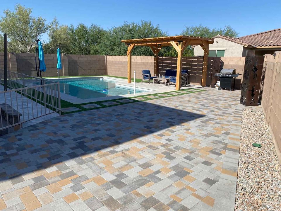 A backyard by Prescott Turf and Pavers features a swimming pool with a safety fence, a pergola with outdoor seating, two blue umbrellas, a barbecue grill, and a paved patio area with stone tiles. Trees and a house complete the serene setting.