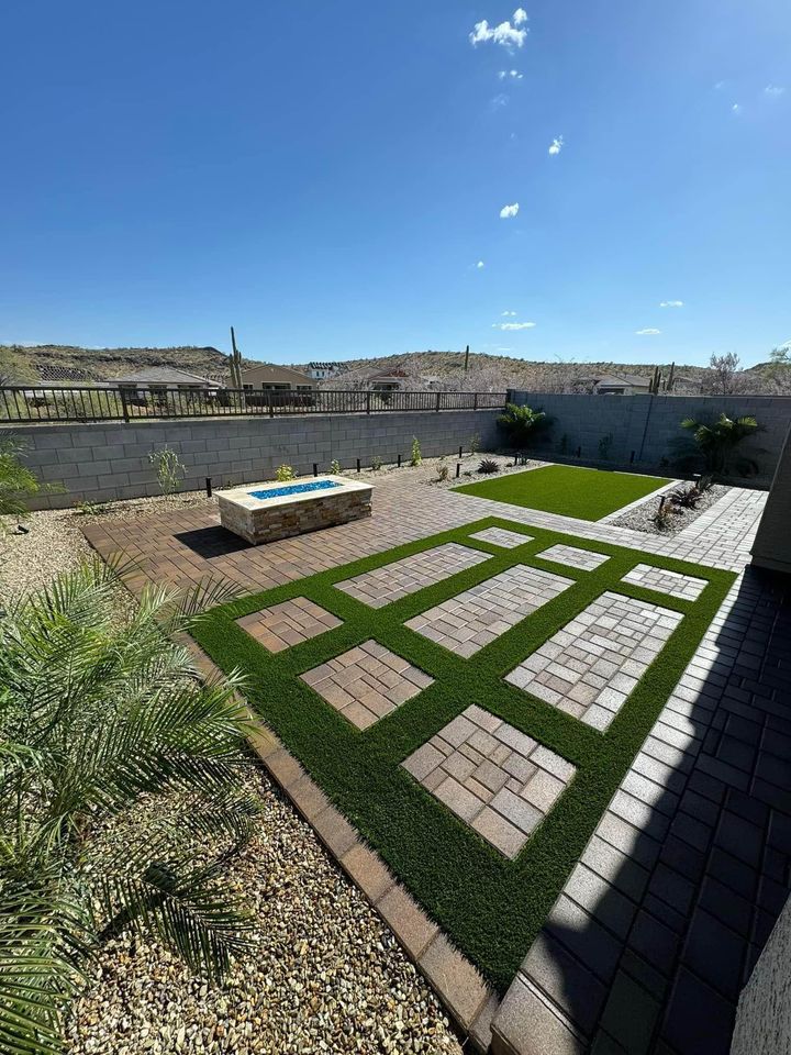 A modern backyard by Prescott Turf and Pavers features geometric patterns of pavers and artificial grass, a stone fire pit, desert landscaping with rocks and cacti, and a block wall fence under a clear blue sky.