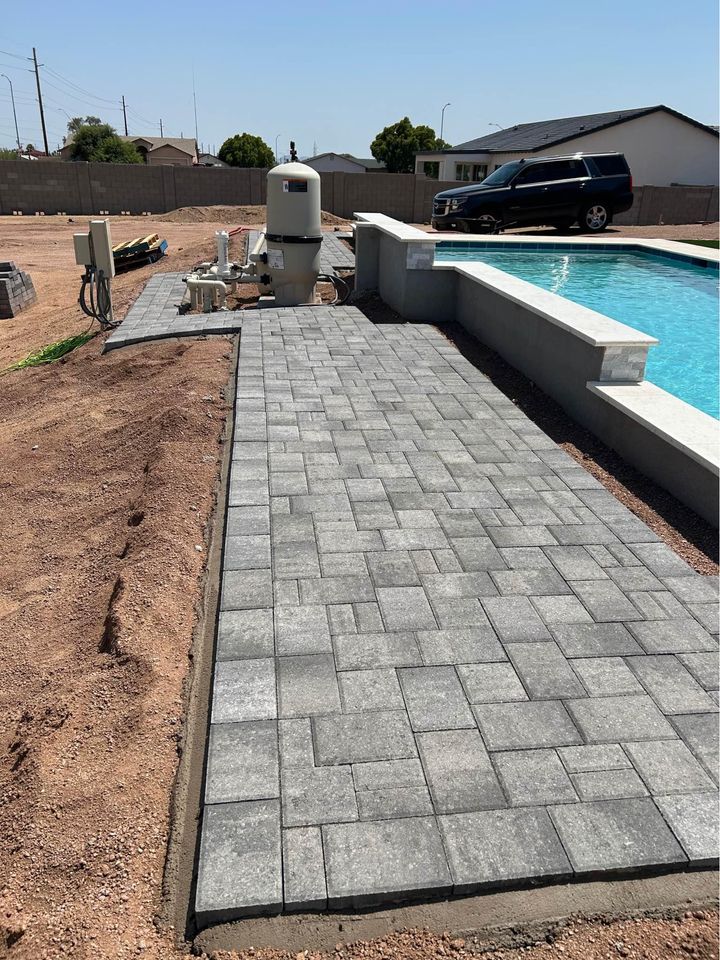 A modern backyard in San Tan Valley, AZ, showcases a rectangular swimming pool with a connected hot tub. The outdoor space is beautifully enhanced with stone tiles and artificial grass. A fence, large house, and cloudy sky create a serene backdrop for this stunning area.