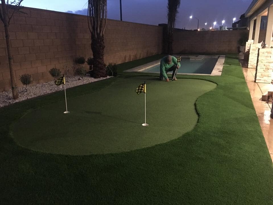 A person installs artificial turf on a backyard putting green with two golf flags, beside a narrow swimming pool and decorative stone landscaping, in the evening.