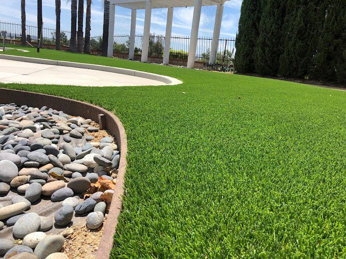 A landscaped yard with neatly trimmed artificial grass, smooth gray river rocks in a bordered area, tall green hedges on the right, and a white pergola with columns in the background under a blue sky.