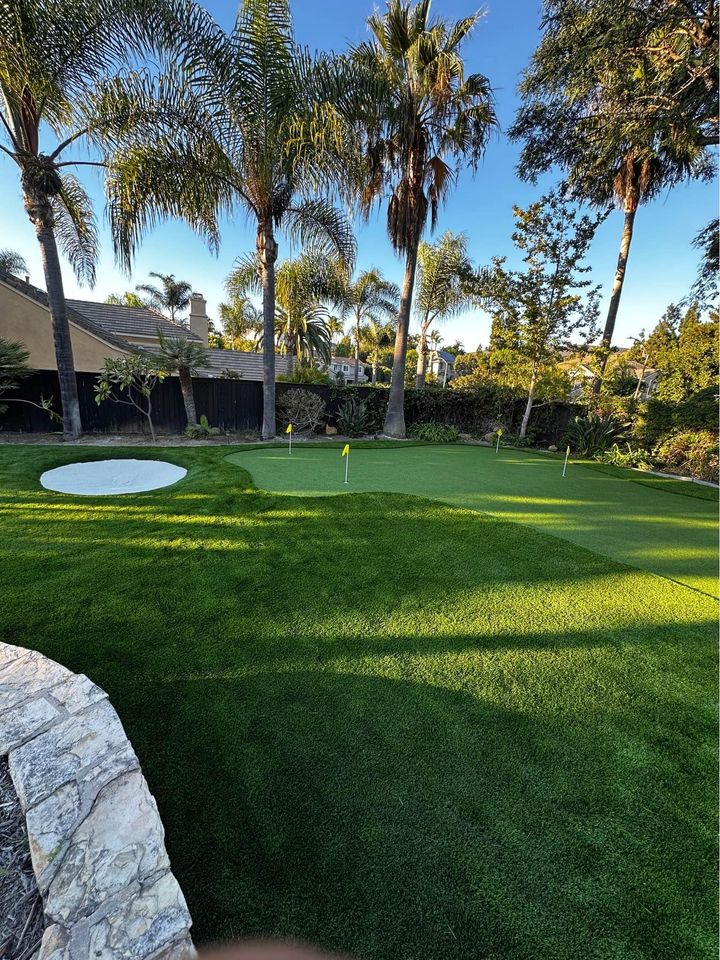 A well-maintained backyard putting green with four flags, surrounded by palm trees and lush landscaping, under a clear blue sky. A small sand bunker sits to the left side of the green.