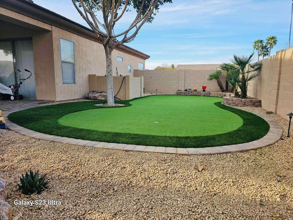 A backyard with artificial grass designed as a putting green, bordered by stones and small desert plants, next to a tan house. A red flag marks a golf hole on the green. A lawnmower is near the window.