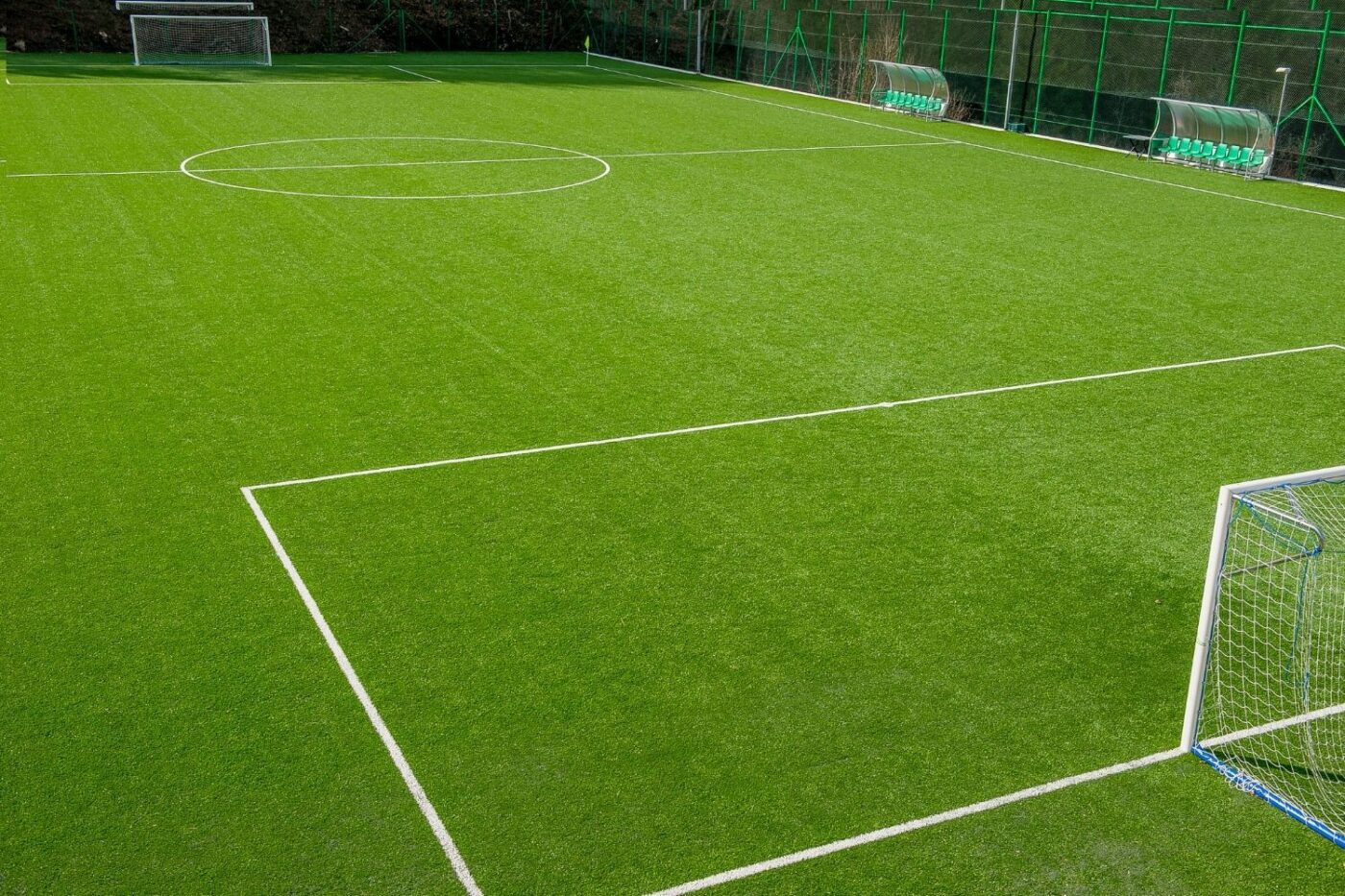 An empty outdoor soccer field with bright green Sports Turf, crisp white boundary lines, two goals, and a row of green benches along the fenced sideline.