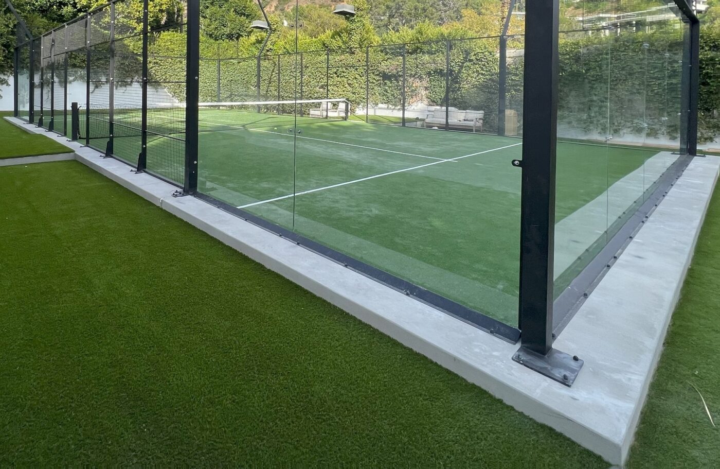 A glass-enclosed outdoor padel court features vibrant green Sports Turf with crisp white lines, surrounded by lush greenery and a bench in the background.