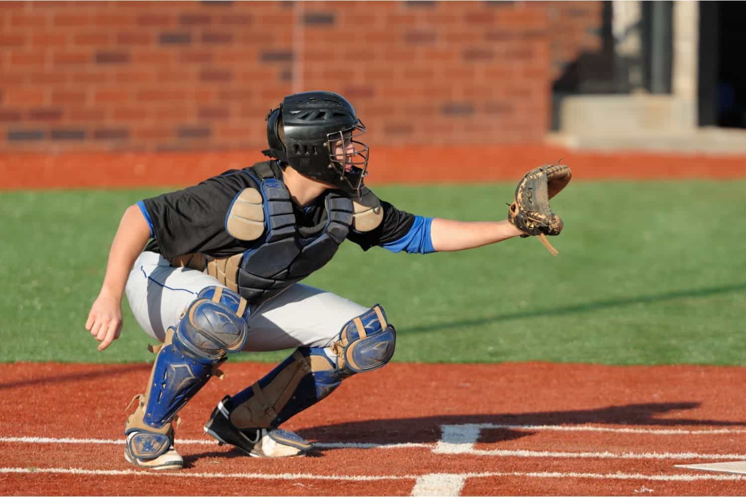 A baseball catcher in full gear crouches behind home plate on the sports turf, wearing a helmet, chest protector, and shin guards, with a glove extended to catch a pitch.
