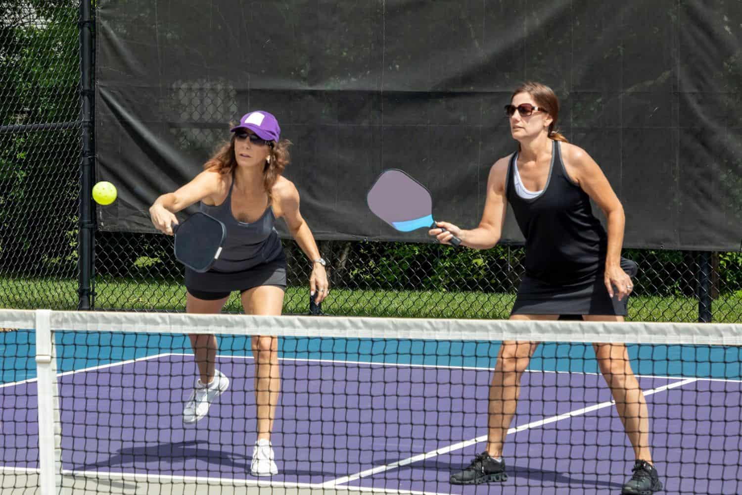 Two women playing pickleball on an outdoor Sports Turf court, both focused on the ball. One woman is preparing to hit it with her paddle while the other stands ready. A net and black fence are in the background.