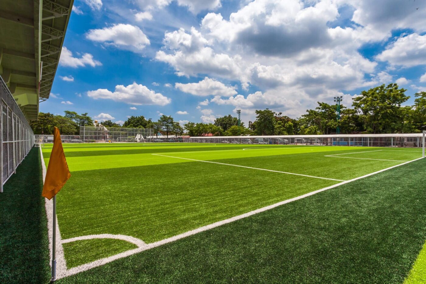 A vibrant green Sports Turf soccer field with clear white boundary lines, an orange corner flag, and a metal fence surrounding the field. The sky above is bright blue with scattered clouds and trees line the background.