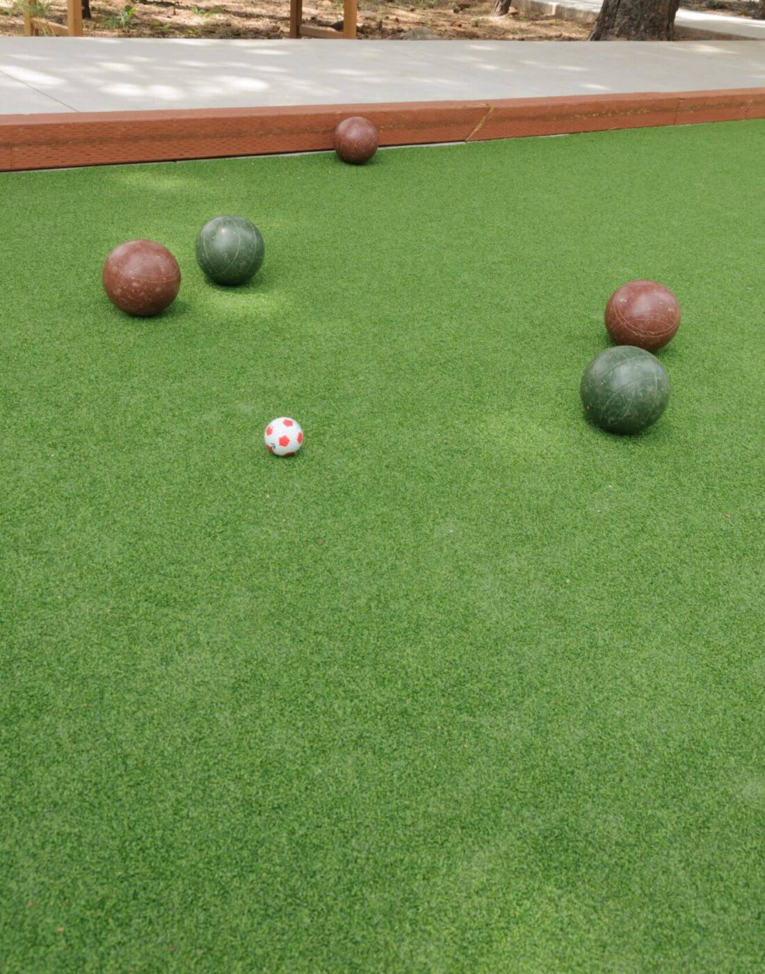Four bocce balls, two green and two brown, are scattered on Sports Turf. A small white and red pallino ball is near the center of the image, with a wooden border lining the court in the background.