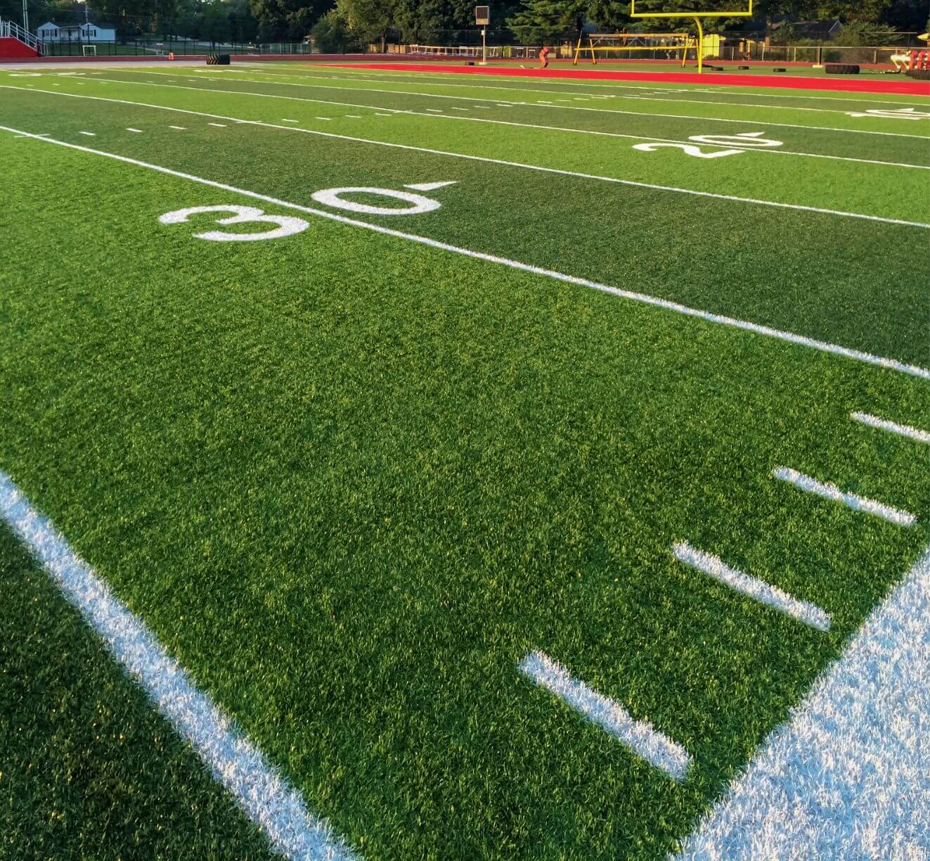A close-up view of a football field shows the 30-yard line and crisp white boundary markings on green sports turf, with goalposts and bleachers visible in the background.